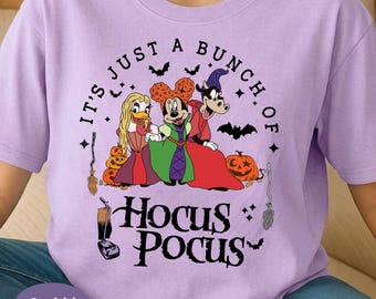 May include: A light purple t-shirt featuring a graphic of three cartoon characters dressed as witches. The text reads IT'S JUST A BUNCH OF HOCUS POCUS. The design includes pumpkins, bats, stars, and two brooms in black ink on the front of the shirt.