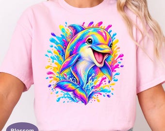 May include: A light pink t-shirt featuring a vibrant, multi-colored graphic of a dolphin leaping out of water. The dolphin is rendered in bright shades of yellow, blue, purple, and pink, surrounded by colorful water splashes and droplets.