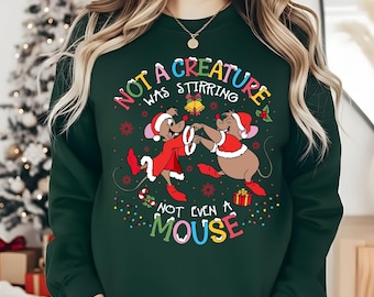 May include: A dark green long-sleeve sweatshirt featuring a graphic of two cartoon mice wearing Santa hats and red coats. The text reads, "NOT A CREATURE WAS STIRRING NOT EVEN A MOUSE," surrounded by colorful holiday lights, bells, and snowflakes.