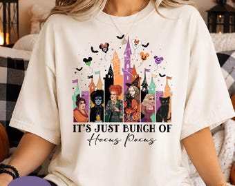 May include: An ivory t-shirt featuring a graphic print of five characters, a black cat, and a castle silhouette. Above the castle are colorful balloons and bats. The text reads IT'S JUST BUNCH OF Hocus Pocus in black, stylized lettering.