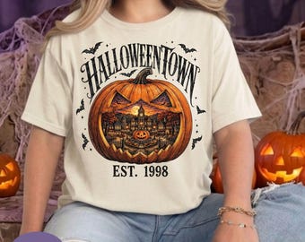 May include: An ivory t-shirt featuring a large orange jack-o'-lantern graphic with a detailed scene of a town inside. The text HALLOWEENTOWN is arched above the pumpkin, and EST. 1998 is printed below. Small black bats are scattered around the design.