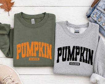 May include: An olive green sweatshirt and a heather gray sweatshirt, both featuring the text PUMPKIN SEASON in a distressed, arched font. The olive sweatshirt has orange text, while the gray sweatshirt has black text, both with a crew neckline.