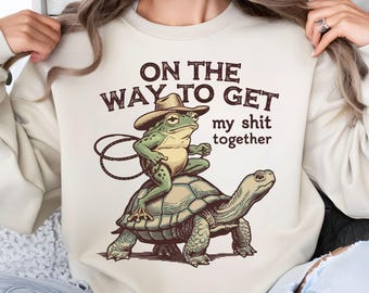 May include: Cream-colored sweatshirt featuring a whimsical illustration. A frog wearing a cowboy hat rides a turtle, with the text "ON THE WAY TO GET my shit together." The design is in shades of green, brown, and cream.