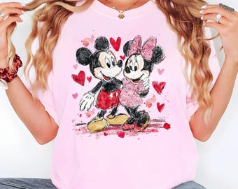 May include: A light pink t-shirt featuring a watercolor illustration of Mickey and Minnie Mouse embracing. Mickey wears red shorts, and Minnie wears a pink dress and bow. Red hearts are scattered around the couple.