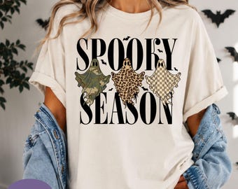 May include: An ivory t-shirt featuring the black text SPOOKY SEASON and three ghost illustrations. The left ghost has a green camouflage pattern, the middle ghost has a brown leopard print, and the right ghost has a tan and white checkerboard pattern.