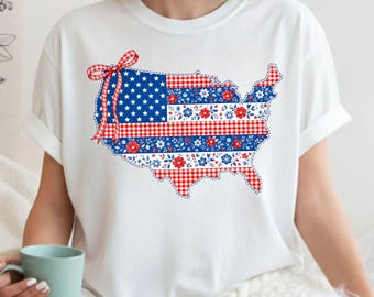 May include: White t-shirt featuring a patriotic design of the United States map. The map is filled with patterns of the American flag, gingham, and floral designs in red, white, and blue. A red and white bow is at the top left.