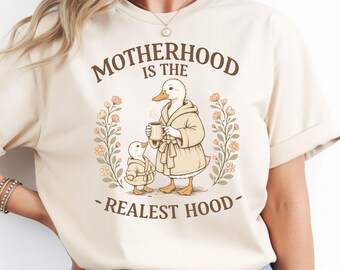 May include: Beige t-shirt with the text "MOTHERHOOD IS THE REALEST HOOD" in brown. The graphic features a mother duck and duckling in bathrobes, holding mugs, surrounded by floral elements. A cute and cozy design.