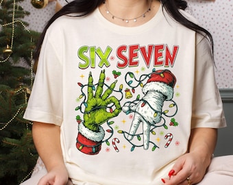 May include: Cream-colored t-shirt with the words "SIX SEVEN" in green and red. The shirt features a Grinch-like hand and a hand in a Santa hat, both adorned with Christmas lights. Festive holiday apparel.