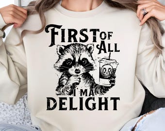 May include: Cream-colored sweatshirt with a black and white graphic of a raccoon holding a coffee cup with a skull design. The text "FIRST OF ALL I'M A DELIGHT" is printed in black, distressed font.