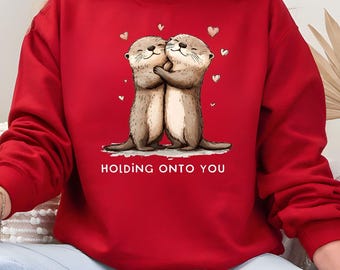 May include: A red sweatshirt featuring a graphic of two hugging otters with hearts. The text "HOLDING ONTO YOU" is printed below the otters. The otters are brown and tan, and the hearts are white.