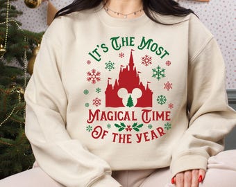 May include: Cream-colored sweatshirt with a festive design. The graphic features a red castle, a green Christmas tree, snowflakes, and the text "It's the Most Magical Time of the Year" in green and red. A holiday-themed apparel item.