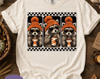 May include: An ivory t-shirt featuring a graphic print of three raccoons wearing orange knit beanies and heart-shaped sunglasses. Each raccoon holds a coffee cup decorated with a pumpkin illustration. The design is framed by black and white checkered borders.