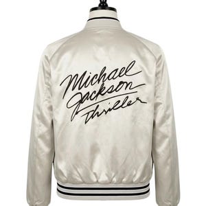 Puede incluir: Chaqueta bomber de satén color crema con detalles a rayas blancas y negras. La parte trasera muestra las palabras "Michael Jackson Thriller" en escritura negra. La chaqueta tiene un estilo bomber clásico con cuello y puños acanalados.