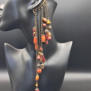 Op de afbeelding: Een ear cuff met meerdere bungelende bronskleurige kettinkjes, versierd met diverse kralen in oranje, rode en koraaltinten, afgewisseld met kleine metalen filigraanaccenten die in een gelaagde, trapsgewijze opstelling naar beneden hangen.