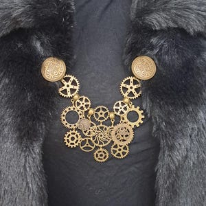 Op de afbeelding: Een steampunk-stijl ketting met een reeks bronzen tandwielen en tandkransen van verschillende formaten. De ketting is versierd met twee ronde, sierlijke bronzen sluitingen. De ketting wordt getoond tegen een zwarte achtergrond en een zwarte imitatiebont kraag.
