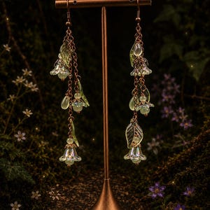 Può includere: Un paio di lunghi orecchini pendenti con catenelle color rame decorate con perline di vetro a forma di fiore verde pallido iridescente e piccoli ciondoli a forma di foglia, sospesi a ganci di metallo color rame su un supporto a T.