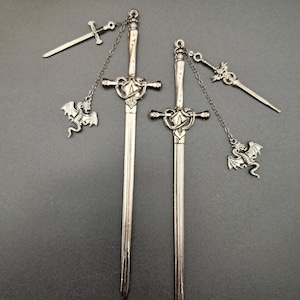 Könnte beinhalten: Ein Paar silberfarbene Ohrringe mit einem langen Schwertanhänger und einem herzförmigen Parierstange. Jeder Ohrring verfügt über eine feine Kette, die das Schwert mit einem kleinen Drachenanhänger und einem weiteren kleinen Schwert verbindet.
