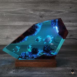 May include: A unique resin lamp featuring a miniature underwater scene with sea turtles, a shipwreck, and coral. The lamp has a wooden base and a geometric shape, with a blue glow illuminating the details.