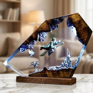 Può includere: Scultura decorativa in resina raffigurante un mago su una scopa volante davanti a un castello. La scultura ha una base e una cornice in legno con dettagli blu e bianchi. Il testo "CapturedLightStudio" è visibile sulla base.