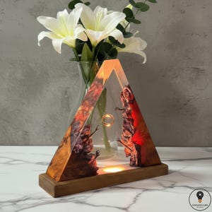 May include: A triangular resin lamp with a wooden base, featuring a gold ring suspended inside. The lamp's interior showcases a textured, reddish-brown design, and the base has a warm, amber glow. White flowers in a glass vase are in the background.