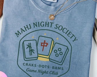 May include: A blue jean Comfort Colors t-shirt with the text "MAHJ NIGHT SOCIETY" and a graphic of three Mahjong tiles. The shirt also features the text "CRAKS DOTS BAMS Game Night Club". A gold necklace with a pendant is draped across the shirt.