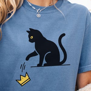 May include: A blue t-shirt featuring a black cat with yellow eyes, pawing at a falling yellow crown. The cat is sitting on a black line. The design is centered on the shirt.