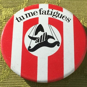 May include: A red and white striped button with the text "tu me fatigues" in black lettering. A black silhouette of a puppet with strings is in the centre of the button.