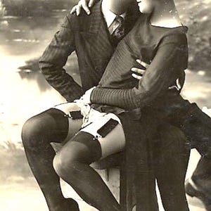Può includere: Una fotografia vintage in bianco e nero che ritrae due persone in abbigliamento formale. Una persona indossa un abito gessato e una cravatta, mentre l'altra indossa un top a maniche lunghe, reggicalze con calze e tacchi scuri, seduta su un blocco.