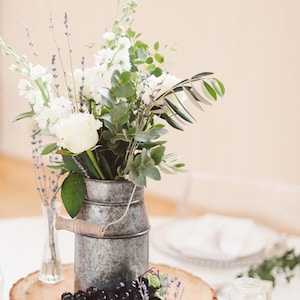May include: A rustic wedding centerpiece featuring a galvanized milk churn filled with white flowers and greenery. The arrangement sits on a wooden slice, adorned with grapes and small vases. The text reads: HOW TO STYLE A MILK CHURN WEDDING CENTREPIECE.