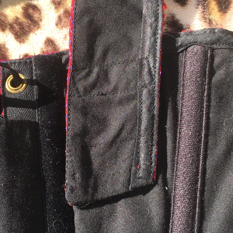 May include: Close-up of a black garment with a red and purple trim. The garment has a hook and loop fastener and a gold-colored grommet. The fabric appears to be a sturdy material, possibly cotton or a blend. The background has a leopard print.