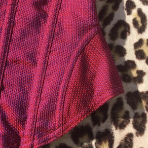 May include: Close-up of a section of a red and purple textured fabric. The fabric has a woven appearance with vertical seams and a curved edge. The background features a leopard print pattern.