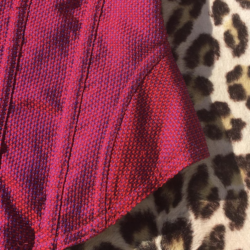 May include: Close-up of a section of a red and purple textured fabric. The fabric has a woven appearance with vertical seams and a curved edge. The background features a leopard print pattern.