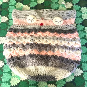 Puede incluir: Un nido de bebé hecho a mano con forma de búho de ganchillo en tonos grises, rosas y blancos. El búho tiene ojos grandes y redondos y un pequeño pico rojo. El cuerpo presenta un patrón texturizado en forma de escamas.