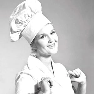 Könnte beinhalten: Eine lächelnde Person in weißer Kochmütze und Uniform. Die Kochmütze ist hoch und plissiert, und die Uniform hat ein doppelreihiges Design. Die Person schaut in die Kamera und lächelt.