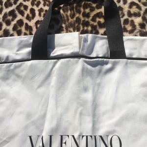 May include: White tote bag with black handles and trim. The word "VALENTINO" is printed in black on the front of the bag. The background is a leopard print pattern.