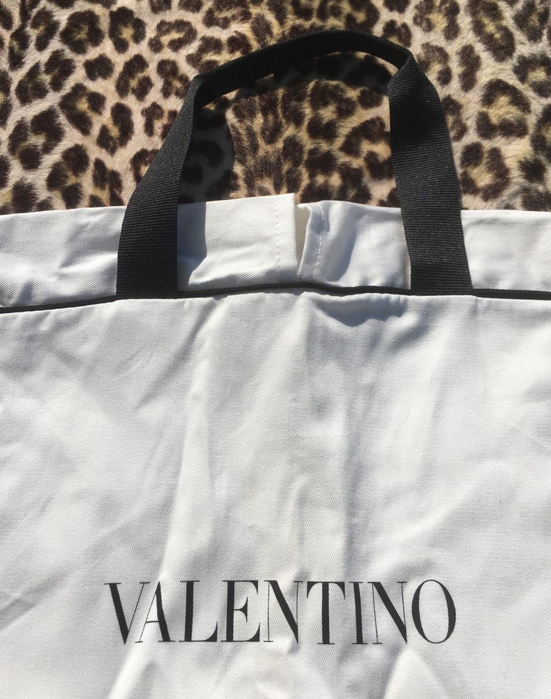 May include: White tote bag with black handles and trim. The word "VALENTINO" is printed in black on the front of the bag. The background is a leopard print pattern.