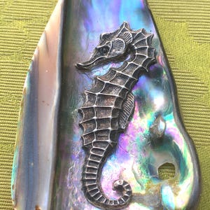 Op de afbeelding: Een hanger met een zeepaardontwerp. Het zeepaardje is een gedetailleerd, zilverkleurig reliëf tegen een glinsterende, iriserende abalone schelp achtergrond. De schelp toont tinten blauw, paars en groen.