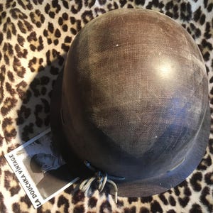 Può includere: Un casco marrone vintage con una superficie strutturata e una corona arrotondata. Il casco ha una tesa e un sottogola. Una piccola etichetta con la scritta "LA DOUCHKA VINTAGE" è visibile. Il casco è su uno sfondo con stampa leopardata.