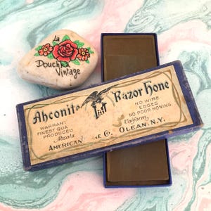 Puede incluir: Piedra vintage con una rosa roja pintada a mano y las palabras "La Douchka Vintage". Una caja azul con la etiqueta "Ahconite Razor Hone" con texto y un águila está abierta, revelando una piedra de afilar marrón. La caja dice "American".
