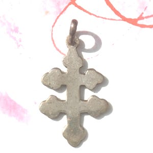 May include: A small, silver-colored pendant in the shape of a double cross. The cross has rounded edges and a loop at the top for a chain. The pendant is likely made of metal and has a slightly aged appearance.