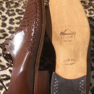 May include: Brown leather dress shoes with woven detailing on the upper. The shoes have a square toe, a block heel, and lace-up closure. The sole is visible, with text that reads "CUIR VERITABLE" and "TRESSE MAIN SUR FORME".