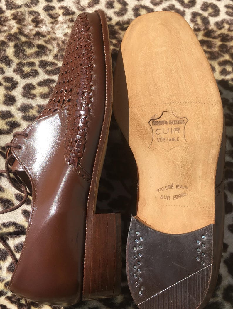 May include: Brown leather dress shoes with woven detailing on the upper. The shoes have a square toe, a block heel, and lace-up closure. The sole is visible, with text that reads "CUIR VERITABLE" and "TRESSE MAIN SUR FORME".