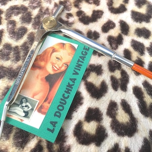 Può includere: Strumento da disegno vintage con corpo in metallo argentato e manico arancione. Un'etichetta verde con la scritta "LA DOUCHKA VINTAGE" e un'immagine pin-up è allegata. Lo sfondo è un motivo leopardato.