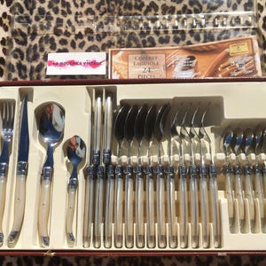 May include: A 24-piece Laguiole flatware set in a brown presentation box. The set includes forks, knives, and spoons with polished metal heads and cream-colored handles. The box lid features the text Coffret Laguiole 24 Pieces.