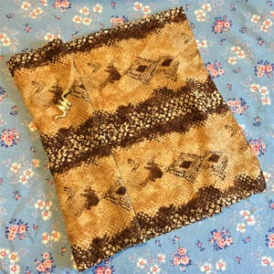 Può includere: Un tessuto con motivo a pelle di serpente marrone e beige con un ciondolo a forma di serpente dorato. Il tessuto è piegato a metà e ha una superficie testurizzata.