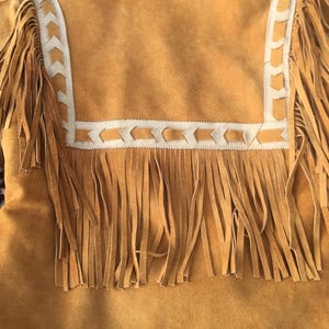 Puede incluir: Chaqueta de ante marr&oacute;n con largos flecos. La chaqueta presenta un ribete decorativo color crema con un patr&oacute;n geom&eacute;trico alrededor del cuello y los hombros. Los flecos cuelgan de las mangas y de la parte inferior de la chaqueta.