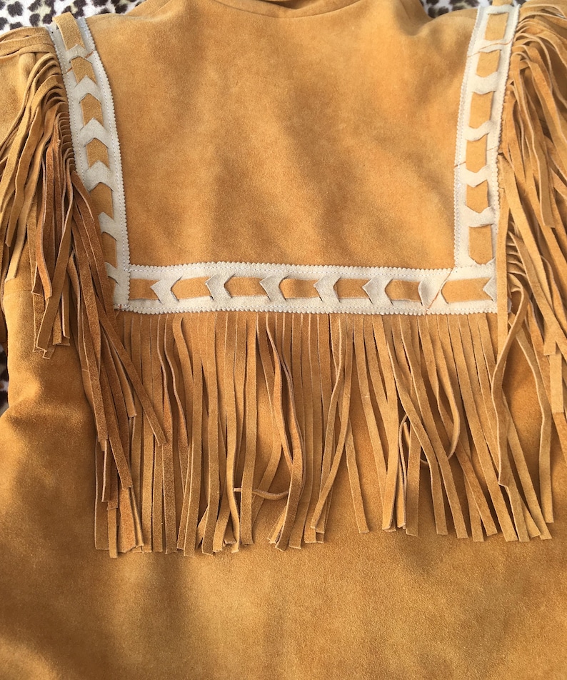 Puede incluir: Chaqueta de ante marr&oacute;n con largos flecos. La chaqueta presenta un ribete decorativo color crema con un patr&oacute;n geom&eacute;trico alrededor del cuello y los hombros. Los flecos cuelgan de las mangas y de la parte inferior de la chaqueta.