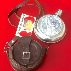 Op de afbeelding: Een vintage zilveren flacon met een leren etui en riem. De flacon heeft een ronde vorm met een zilveren dop. De bruine leren etui heeft een gespsluiting. Een label met de tekst "LA DOUCHKA VINTAGE" is ook aanwezig.