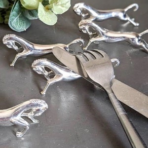 Könnte beinhalten: Ein Set aus vier pferdeförmigen Messerbänken aus silberfarbenem Metall. Jedes Stück zeigt eine stilisierte Pferdefigur in galoppierender Pose, die als Ablage für Besteck dient. Eine Edelstahlgabel liegt quer über einer der Pferdefiguren.