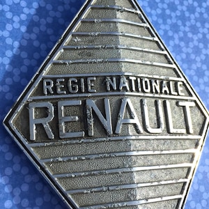 Può includere: Un emblema argentato a forma di diamante con la scritta "REGIE NATIONALE RENAULT". L'emblema ha una superficie strutturata e un design geometrico, probabilmente un distintivo o un logo automobilistico.